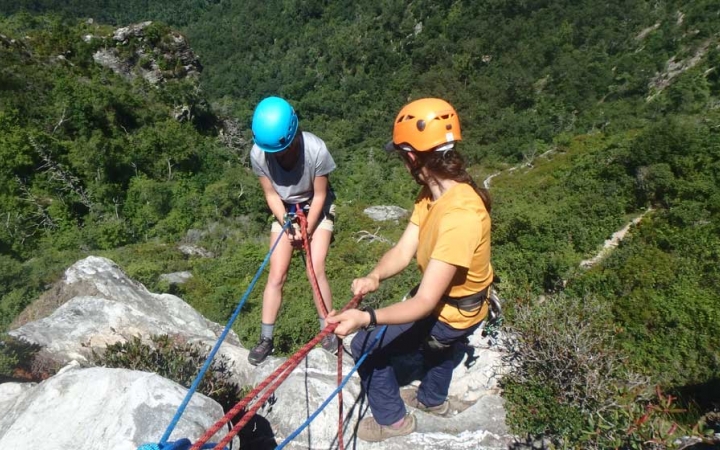 Two people wearing safety gear are secured by ropes near the edge of a cliff. One person appears to be an instructor, giving direction to the other person. Below them is a green wooded area. 